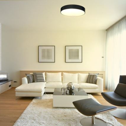 Svetlo svieti v moderne zariadenej obývacej izbe. Biela rohová sedačka je umiestnená na svetlom koberci pred bielym konferenčným stolíkom. V pozadí sú dva obrázky a tmavé kožené kreslo.