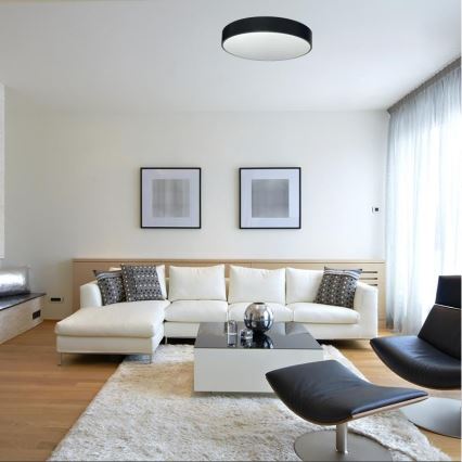 Stropné svietidlo svieti v moderne zariadenej obývacej izbe. Biela rohová sedačka a konferenčný stolík sú umiestnené na svetlom koberci. V pozadí sú dva obrazy a svetlé závesy.