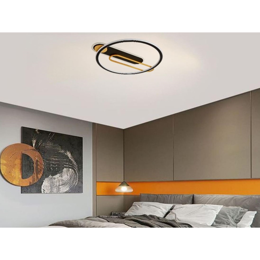 Svietidlo s kruhovým tvarom a čiernymi a zlatými prvkami je pripevnené k stropu. Svietidlo osvetľuje spálňu s posteľou a obrazom na stene.