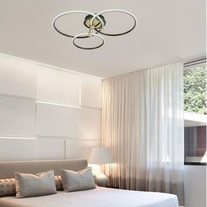 Svietidlo s tromi prepletenými kruhovými prvkami je pripevnené k stropu. Svietidlo je v spálni s posteľou a bielymi závesmi.