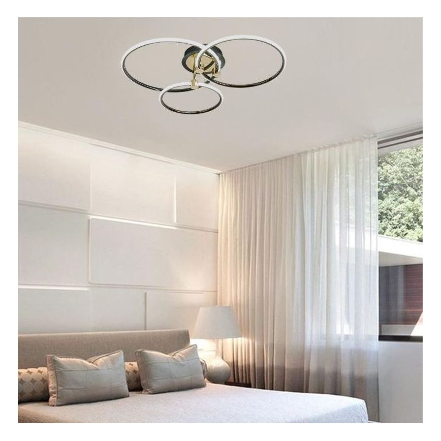 Svietidlo s tromi prepletenými kruhovými prvkami je pripevnené k stropu. Svietidlo je v spálni s posteľou a bielymi závesmi.