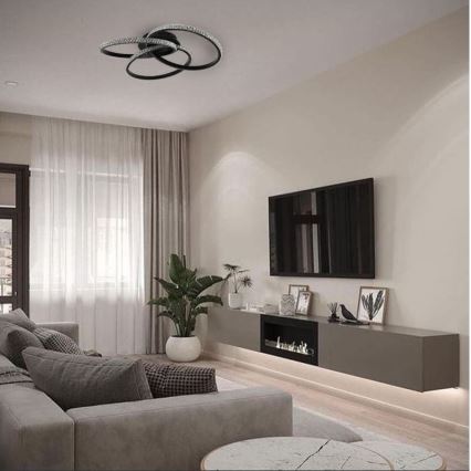 Svietidlo s prepletenými kruhmi a kryštálmi je pripevnené na strope. Svietidlo osvetľuje obývaciu izbu s pohovkou, televízorom a dekoráciami.