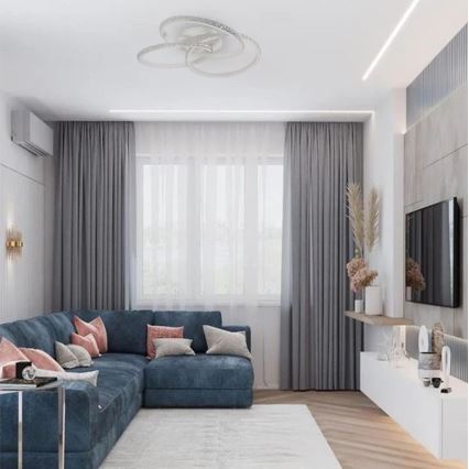 Stropné svietidlo s troma prepletenými kruhmi osvetľuje modernú obývačku. Svietidlo je umiestnené na strope nad rohovou sedačkou, ktorá je doplnená šedými závesmi a televízorom na stene.