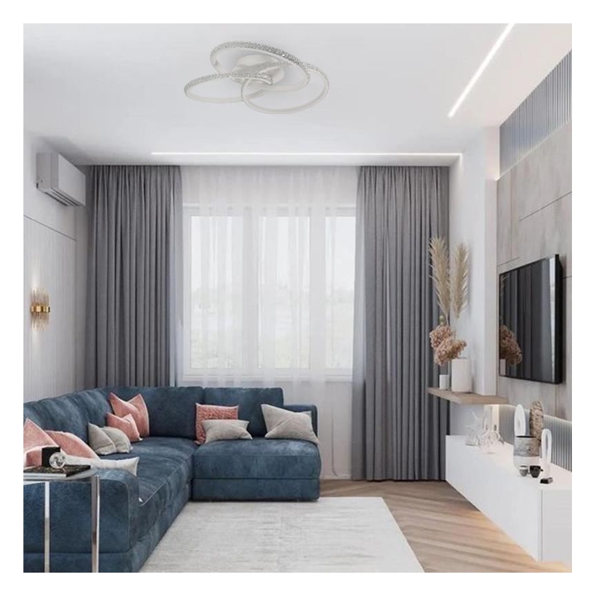 Stropné svietidlo s troma prepletenými kruhmi osvetľuje modernú obývačku. Svietidlo je umiestnené na strope nad rohovou sedačkou, ktorá je doplnená šedými závesmi a televízorom na stene.