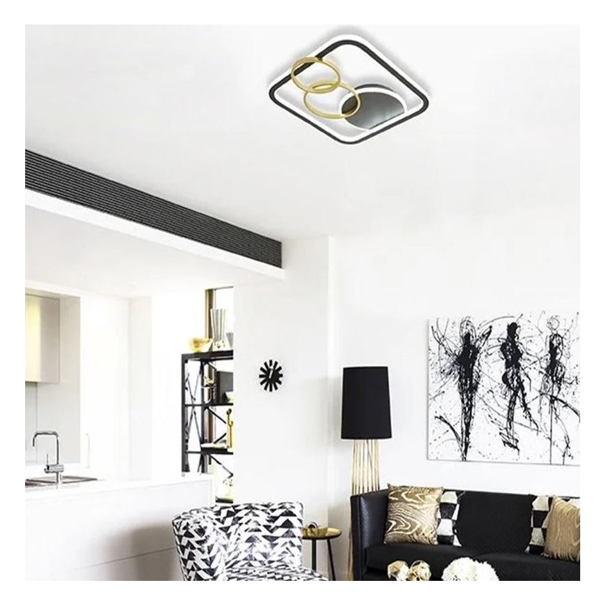 Stropné svietidlo s bielym a zlatým rámom a čiernymi kruhmi je osvetlené. Nachádza sa v modernej obývacej izbe s čierno-bielym nábytkom a doplnkami.