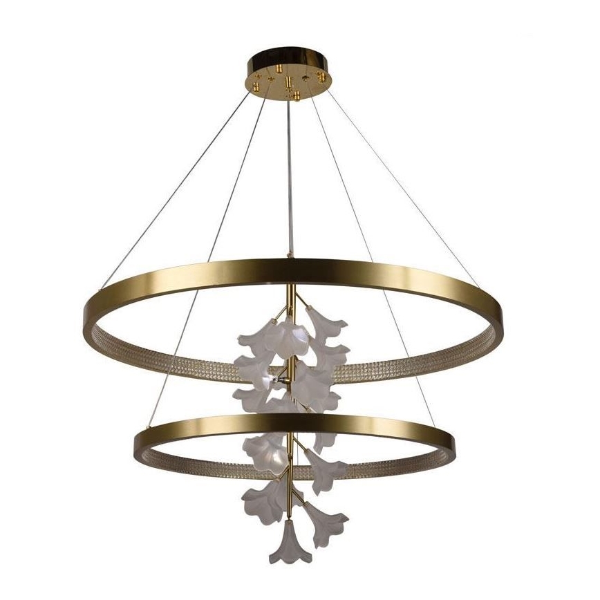 Dvojité zlaté kruhové svietidlo s viacerými bielymi kvetmi visiacimi v strede, osvetľuje priestor. Svietidlo je zavesené zo stropu pomocou tenkých káblov.