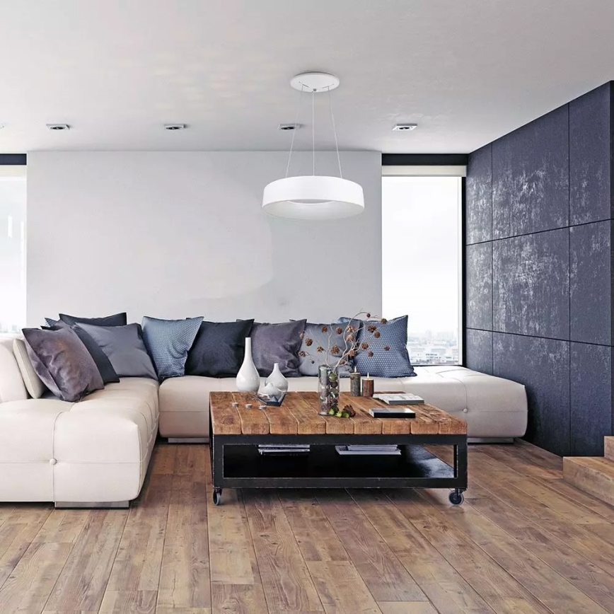 Závesné svietidlo svieti v obývacej izbe. Svietidlo je okrúhle, biele a visí nad rohovou sedačkou a dreveným konferenčným stolíkom. Obývačka má drevenú podlahu a tmavé steny.