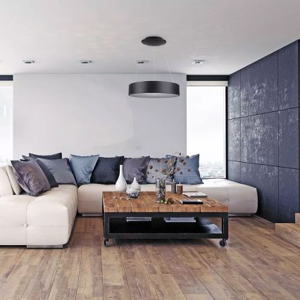 Stropné svietidlo visí v modernom obývacej izbe. Svietidlo osvetľuje bielu rohovú sedačku s vankúšmi a drevený konferenčný stolík. Izba má drevenú podlahu a tmavú stenu.