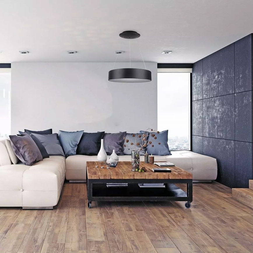 Stropné svietidlo visí v modernom obývacej izbe. Svietidlo osvetľuje bielu rohovú sedačku s vankúšmi a drevený konferenčný stolík. Izba má drevenú podlahu a tmavú stenu.