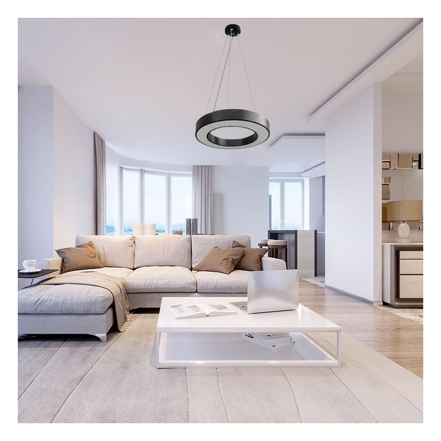 Kruhový luster visí zo stropu v moderne zariadenej obývacej izbe. Luster osvetľuje svetlú sedačku v tvare L a biely konferenčný stolík s notebookom. Izba má veľké okná a svetlé steny.