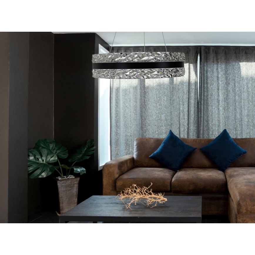 Okrúhle závesné svietidlo s kryštálovým a čiernym prvkom visí v obývacej izbe nad hnedou koženou pohovkou s tmavomodrými vankúšmi. V miestnosti sa nachádza aj konferenčný stolík a zelená rastlina v kvetináči.