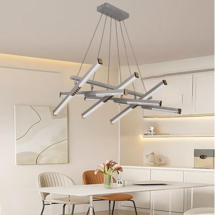 Moderná strieborná LED lustre visí nad jedálenským stolom v minimalistickej jedálni. Lustre pozostáva z viacerých rovnobežných svetelných ramien usporiadaných do asymetrického vzoru. Okolité prostredie tvorí svetlý nábytok a neutrálne steny.