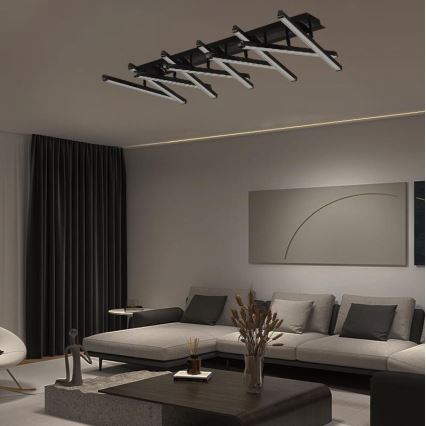 Dlhá čierna stropná lampa s viacerými paralelnými svetlami osvetľuje modernú obývaciu izbu s rohovou sedačkou, konferenčným stolíkom a doplnkami.