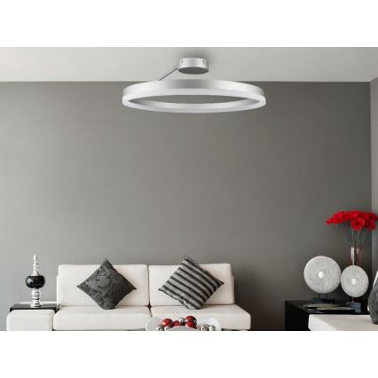 Stropné svietidlo v tvare kruhu svieti v obývacej izbe. Svietidlo je strieborné, pripevnené k stropu a osvetľuje bielu sedačku s vankúšmi a okolité nábytkové doplnky.