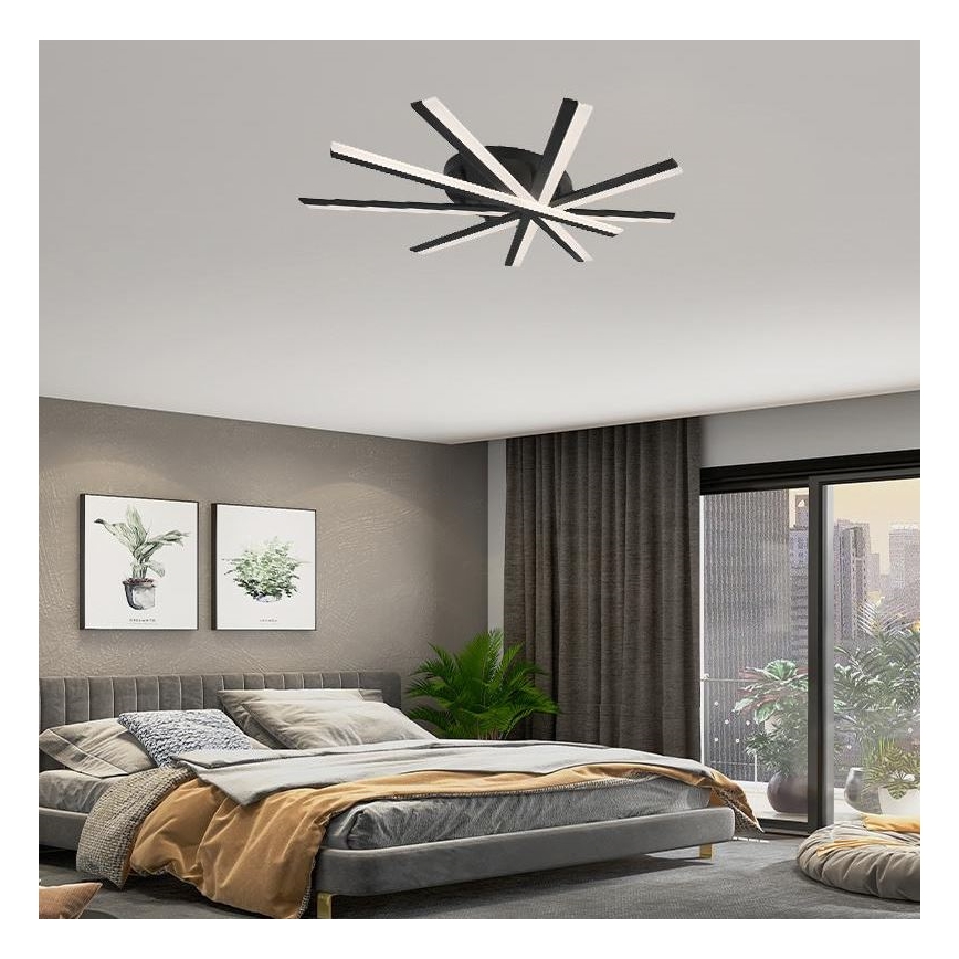 Stropné svietidlo s viacerými ramenami v tvare hviezdice svieti v spálni. Svietidlo je čierne s bielymi LED diódami. Spálňa obsahuje posteľ, závesy a rastliny.