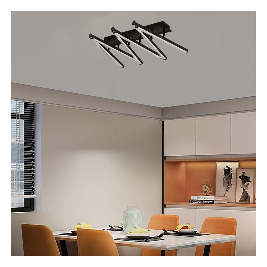 Moderné stropné svietidlo s viacerými ramenami a lineárnymi svetlami svieti v jedálni. Svietidlo je pripevnené k stropu nad obdĺžnikovým jedálenským stolom so stoličkami. V pozadí sú viditeľné kuchynské skrinky a okno so žalúziami.