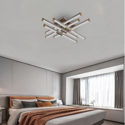 Svietidlo s viacerými paralelnými ramenami svieti v spálni. Ramená sú strieborné s bronzovými koncovkami. Spálňa obsahuje posteľ s oranžovou prikrývkou a šedými vankúšmi. Okno je za posteľou, za ním sú závesy.