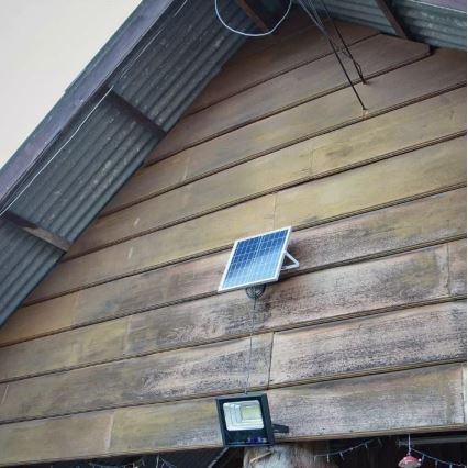 Solárny panel nabíja vonkajšie LED svetlo. Svetlo je pripevnené na drevenú stenu budovy s plechovou strechou.