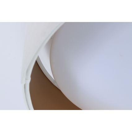 Biela lampa s guľovitým tienidlom sa nachádza na svetlom pozadí. Tienidlo je čiastočne prekryté bielym, zakriveným prvkom s hnedým vnútorným lemovaním.