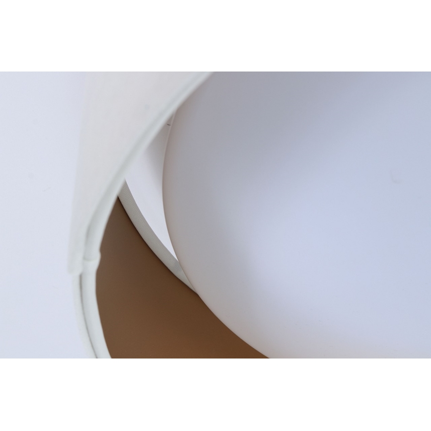 Biela lampa s guľovitým tienidlom sa nachádza na svetlom pozadí. Tienidlo je čiastočne prekryté bielym, zakriveným prvkom s hnedým vnútorným lemovaním.