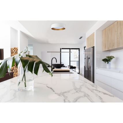 Svietidlo svieti v modernej kuchyni. Svietidlo je biele s zlatým okrajom, umiestnené na strope. Kuchyňa obsahuje kuchynskú linku z bieleho mramoru, čiernu kuchynskú batériu a chladničku.