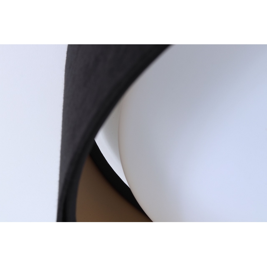 Svietidlo s čiernym tienidlom obopínajúce biely difúzor. Tienidlo čiastočne zakrýva difúzor. Svetlé pozadie.