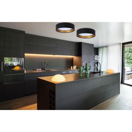 Dve kruhové stropné svietidlá svietia v tmavej kuchyni. Svietidlá sú čierne s vnútorným zlatým okrajom. Kuchynská linka a ostrovček sú tmavosivé.