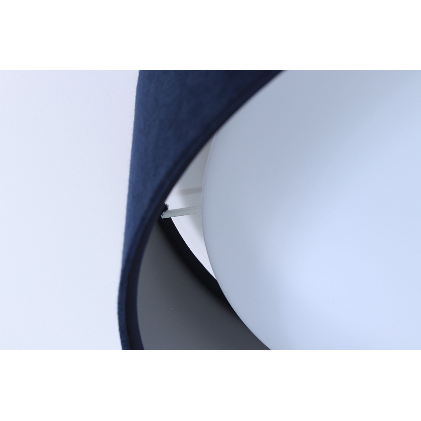 Svietidlo s tmavomodrým textilným tienidlom čiastočne zakrýva bielu guľovú časť. Tienidlo je pripevnené k guli. Svetlý pozadia.