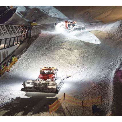 Dva snežné stroje upravujú zjazdovku v noci. Jeden stroj je v popredí, druhý vzadu. Osvetlenie osvetľuje sneh a okolie zjazdovky.