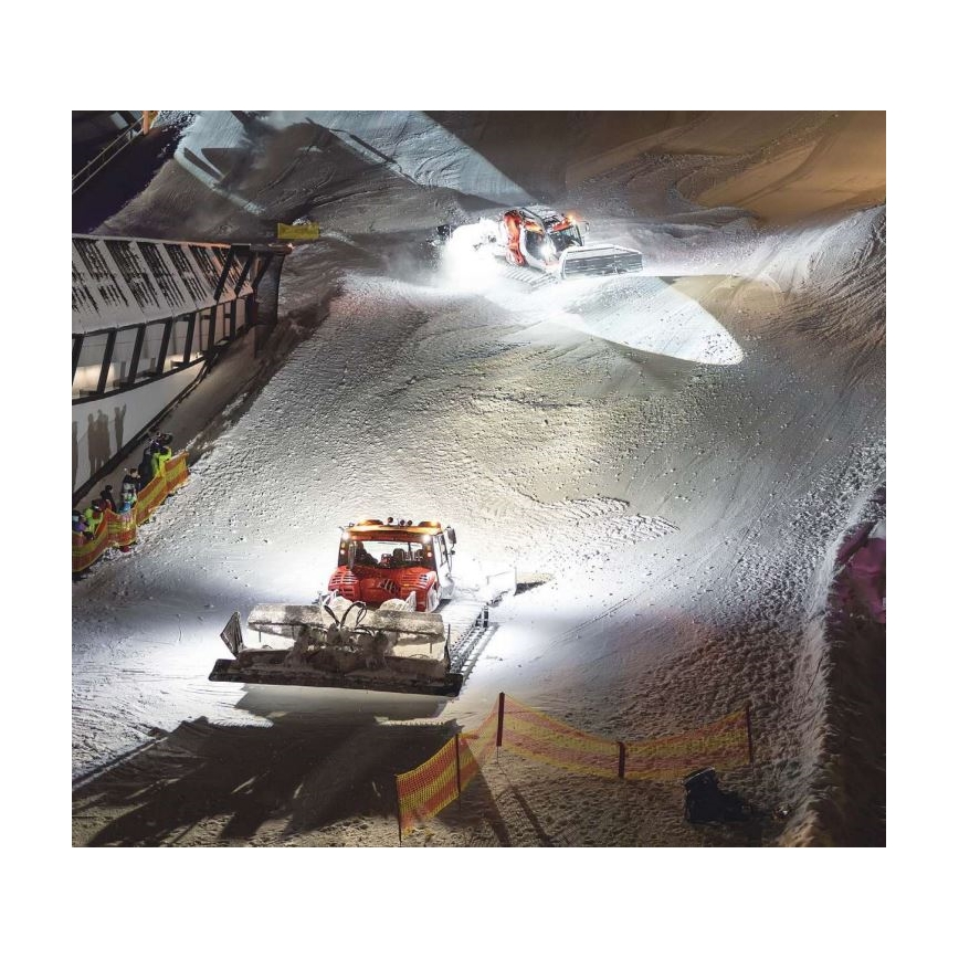 Dva snežné stroje upravujú zjazdovku v noci. Jeden stroj je v popredí, druhý vzadu. Osvetlenie osvetľuje sneh a okolie zjazdovky.