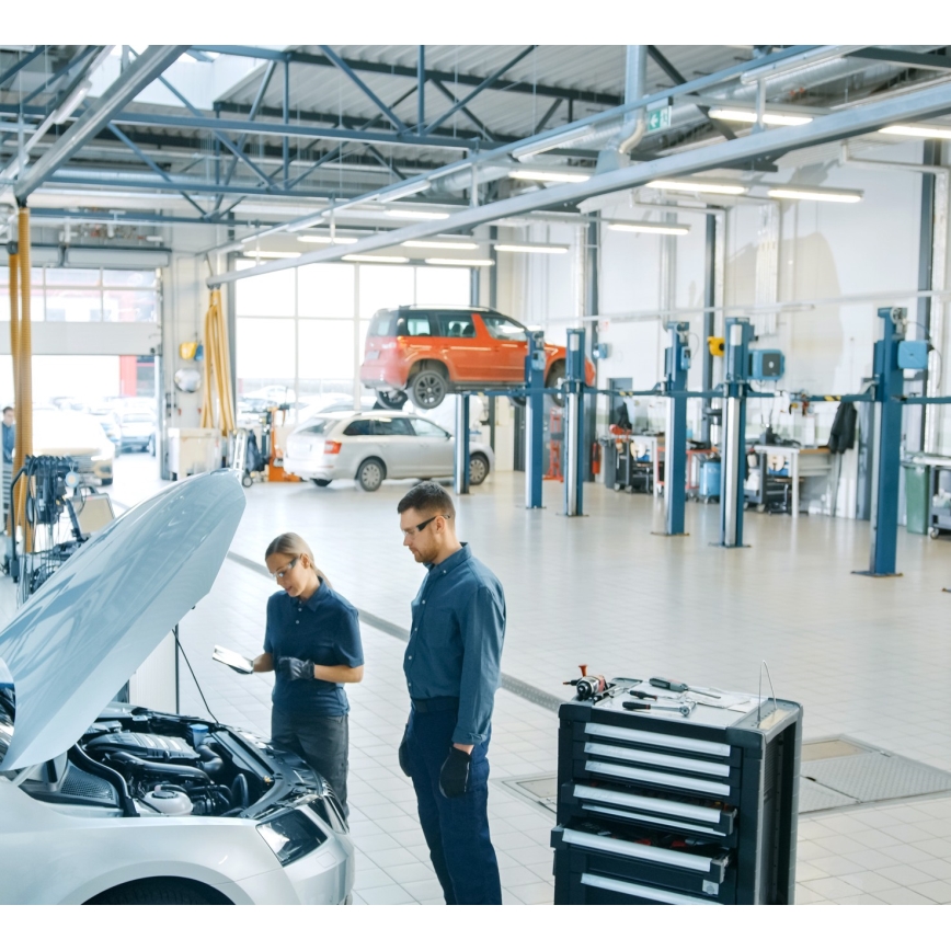 Mechanici kontrolujú motor automobilu. Dvíhacie zariadenia a náradie sú v pozadí veľkej autoservisu.