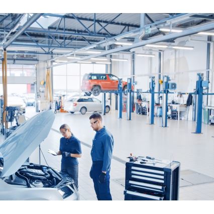 Dva mechanici kontrolujú motor auta s otvorenou kapotou v autoservise. Jeden drží tablet. V pozadí sú ďalšie autá a zdvíhacie plošiny.