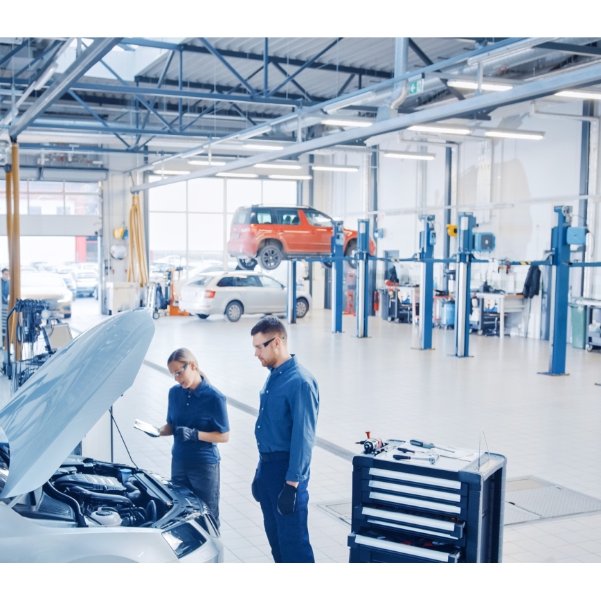 Dva mechanici kontrolujú motor auta s otvorenou kapotou v autoservise. Jeden drží tablet. V pozadí sú ďalšie autá a zdvíhacie plošiny.