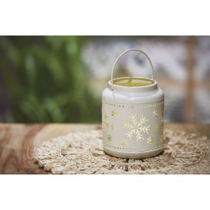 Kovová lucerna s vyrezávanými snehovými vločkami stojí na jutovom obrúsku na drevenom stole. Lucerna má rúčku a vnútri svieti svetlo. Pozadie tvorí rozmazané zelené lístie.
