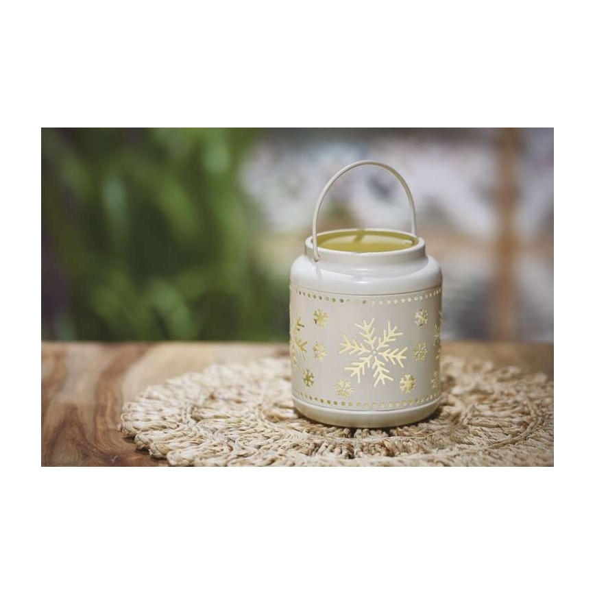 Kovová lucerna s vyrezávanými snehovými vločkami stojí na jutovom obrúsku na drevenom stole. Lucerna má rúčku a vnútri svieti svetlo. Pozadie tvorí rozmazané zelené lístie.