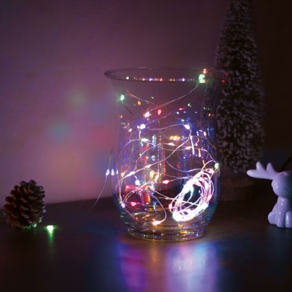 Sklenená váza obsahuje svetielka s farebnými LED diódami. Svetielka sú rozmiestnené vo vnútri vázy, ktorá stojí na tmavom povrchu. V pozadí je umelé vianočné stromček a keramický sobiček.