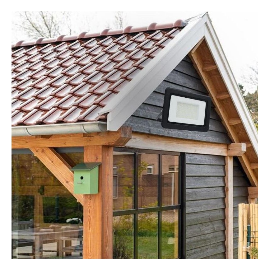 Svetlomét je pripevnený k tmavosivej drevenej stene domu s červenohnedou škridľovou strechou. Na stene je tiež okno a vtáčia búdka.