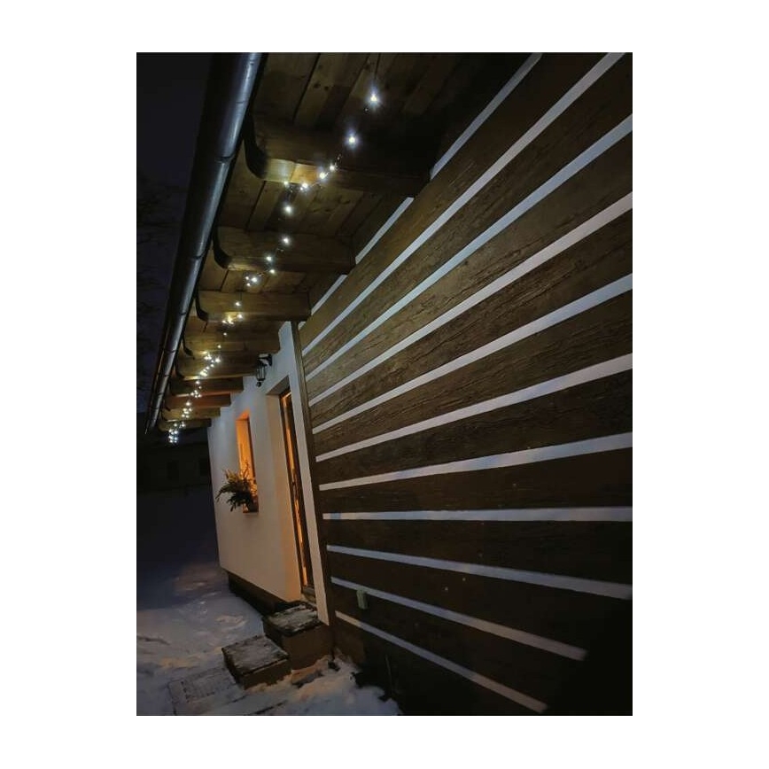 Svetelná reťaz svieti pod strechou domu. Reťaz je umiestnená na drevenom podhľade a osvetľuje stenu z horizontálne kladených drevených dosiek so svetlými medzerami. Sneh pokrýva zem pred domom.