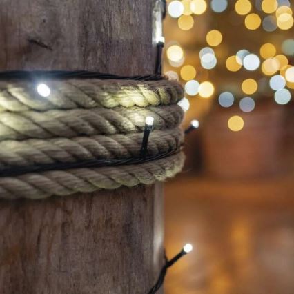 Elektrický svetelný veniec s bielymi LED diódami je omotaný okolo hrubého lana na drevenom stĺpe. V pozadí je rozostrený vianočný stromček s rozžiarenými svetlami.