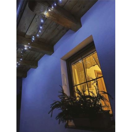 Svetelná reťaz s malými LED diódami je pripevnená pod dreveným stropom. Svetlá osvetľujú modrú stenu a okno s osvetlenou izbou za ním. Pred oknom je kvetináč s rastlinou.