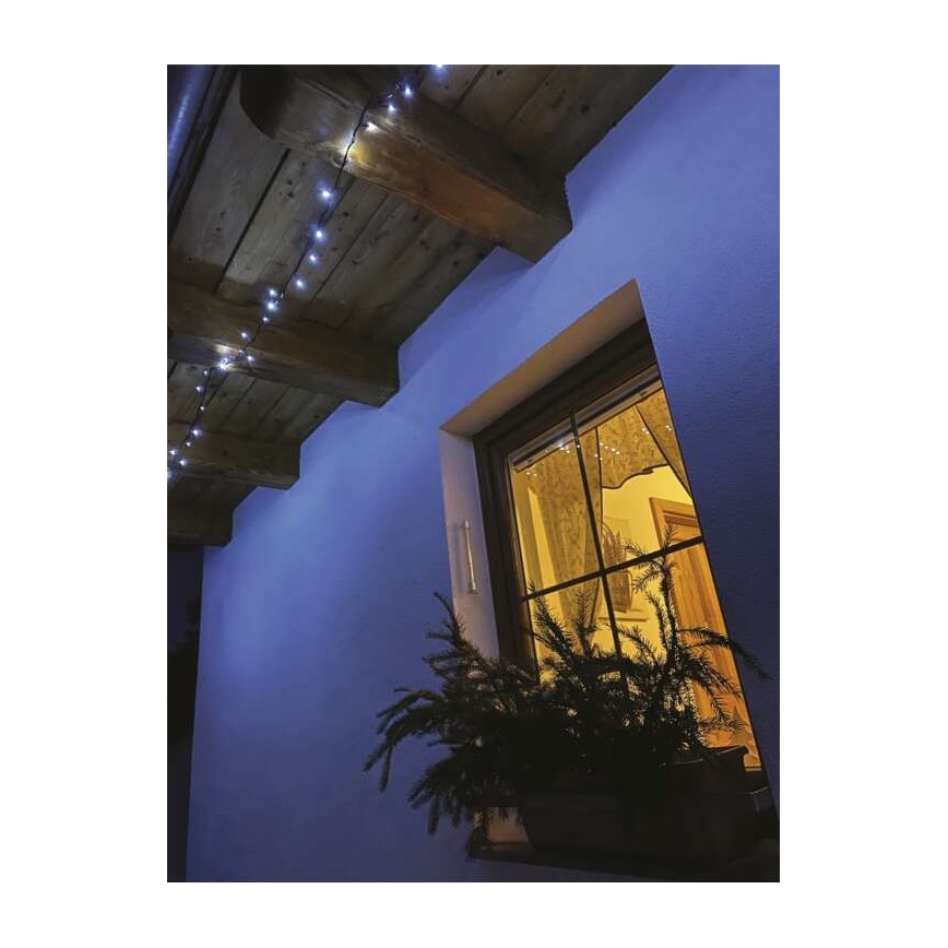 Svetelná reťaz s malými LED diódami je pripevnená pod dreveným stropom. Svetlá osvetľujú modrú stenu a okno s osvetlenou izbou za ním. Pred oknom je kvetináč s rastlinou.