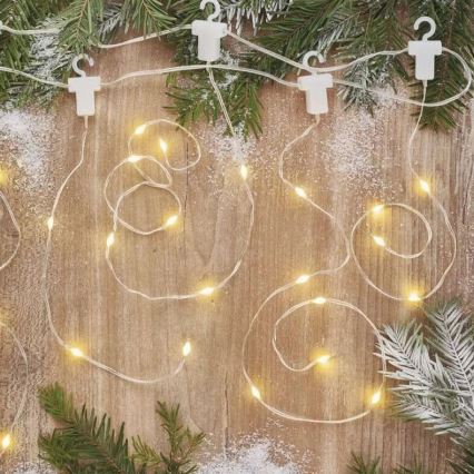 Svetlený elektrický veniec s malými žiarovkami je umiestnený v špirálových závitoch na drevenom povrchu. Veniec je pripevnený k povrchu bielymi sponkami. V okolí sú roztrúsené vetvičky ihličnatého stromu a umelý sneh.