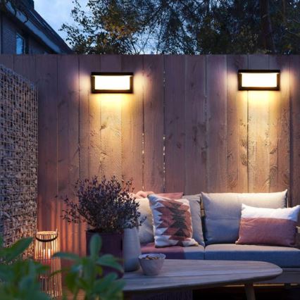 Dve vonkajšie nástenné svietidlá svietia na drevený plot. Pred plotom sa nachádza sedacia súprava s vankúšmi a konferenčný stolík. Atmosféra je večerná, vonkajšie prostredie.