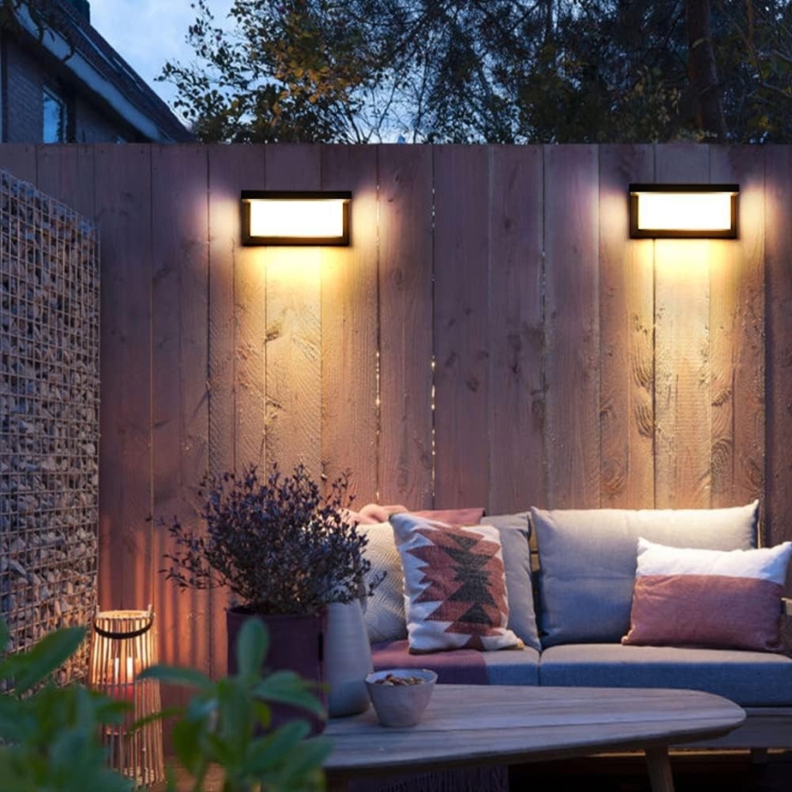 Dve vonkajšie nástenné svietidlá svietia na drevený plot. Pred plotom sa nachádza sedacia súprava s vankúšmi a konferenčný stolík. Atmosféra je večerná, vonkajšie prostredie.