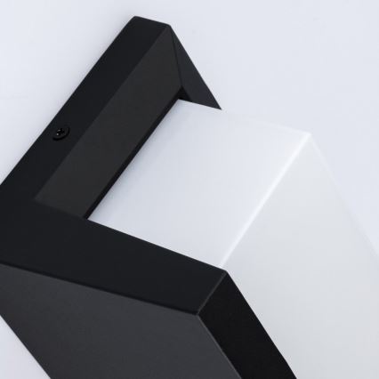 Čierno-biela nástenná lampa s hranatým dizajnom je pripevnená na stene. Lampa je umiestnená v pravom hornom rohu. Viditeľné je pripojenie kábla.