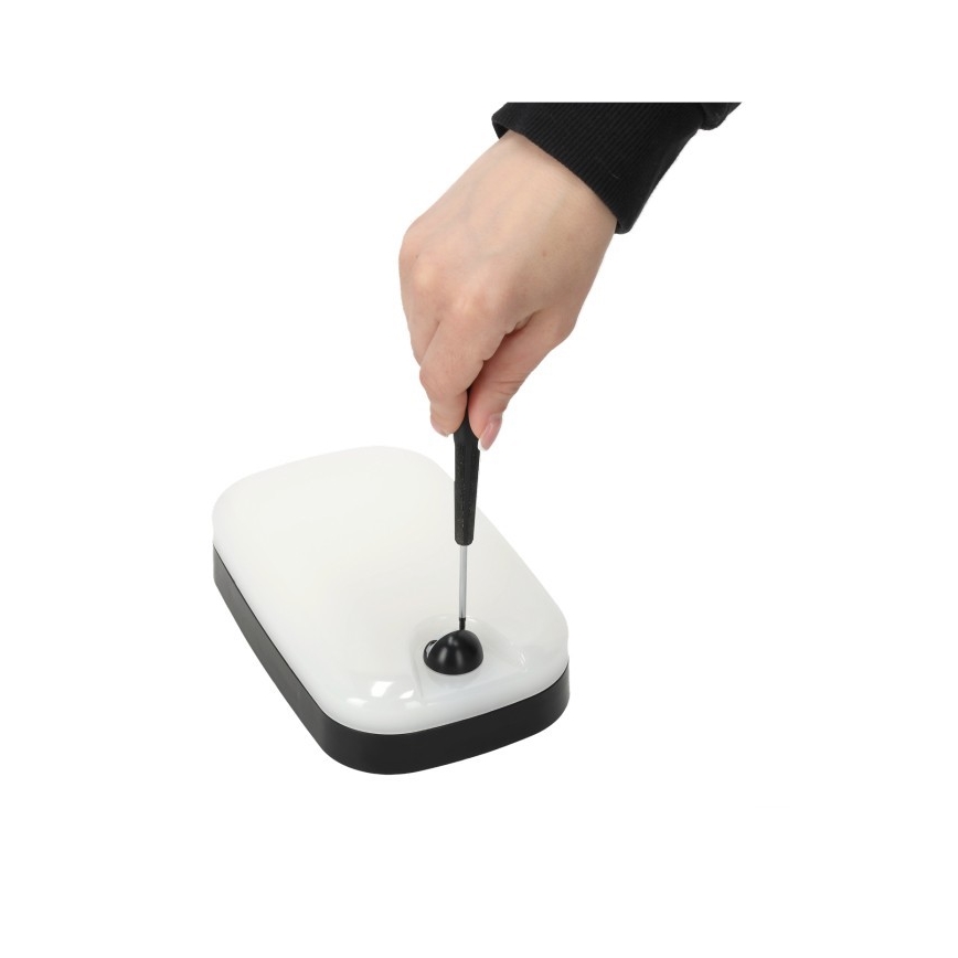 Ruka drží skrutkovač a otáča ním malý čierny diel na obdĺžnikovom bielom svietidle s čiernym rámom. Svietidlo leží na bielom pozadí.