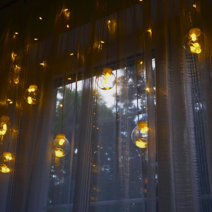Svetielka visia pred oknom so závesmi. Svietia žltým svetlom. Za oknom sa nachádza zeleň.