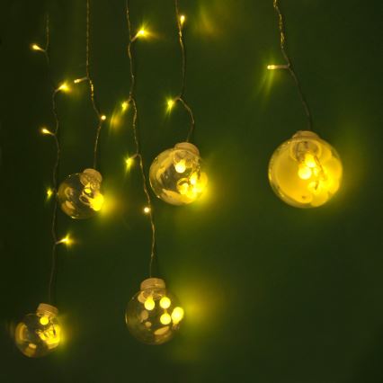 Svetlá vo forme guľôčok s malými žiarovkami vo vnútri visia na tenkých kábloch pred tmavozeleným pozadím. Žiarovky svietia žltým svetlom.