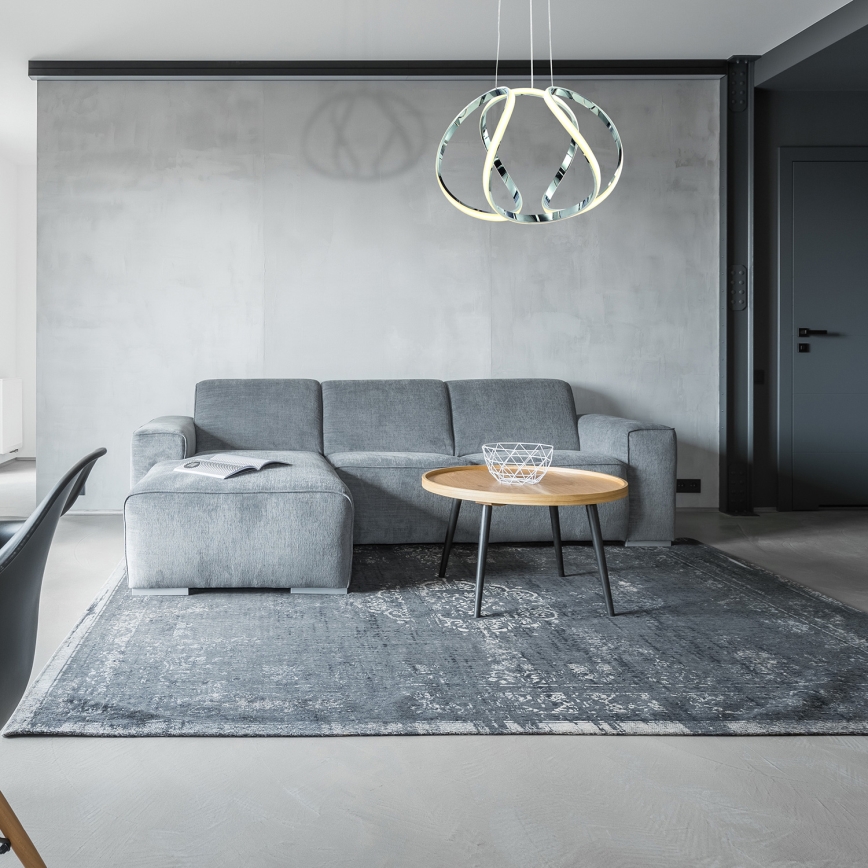 Svietidlo s prepletenými kruhmi visí v modernej obývacej izbe nad televízorom a krbom. Čierna sedačka a konferenčný stolík s ovocím sú súčasťou interiéru.