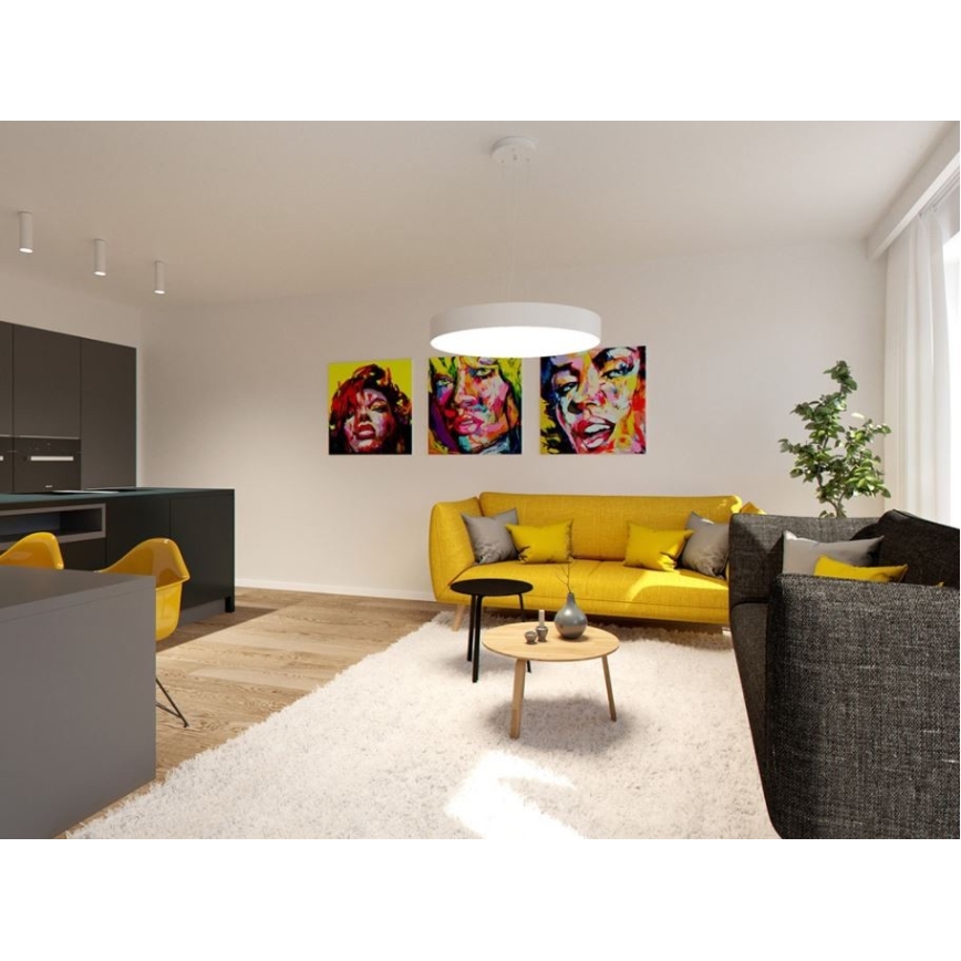 Žltá sedačka stojí v obývacej izbe s drevenou podlahou a bielym kobercom. Na stene visia tri obrazy ženských portrétov v expresívnom štýle. V pozadí je tmavá kuchynská linka.